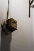 A close-up view of a worn-out, wall-mounted electrical switch with visible dirt and signs of aging. The switch is beige in color, and the wall around it is white with some discoloration.