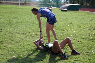 Two athletes are on a grassy field. One athlete is standing and offering a hand to another athlete who is lying on the ground. Both are wearing athletic gear, and it appears to be a moment of sportsmanship or assistance.