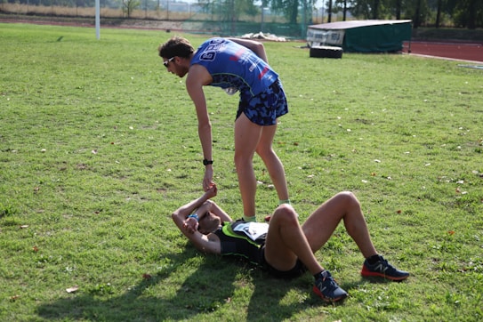 Two athletes are on a grassy field. One athlete is standing and offering a hand to another athlete who is lying on the ground. Both are wearing athletic gear, and it appears to be a moment of sportsmanship or assistance.