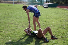 Two athletes are on a grassy field. One athlete is standing and offering a hand to another athlete who is lying on the ground. Both are wearing athletic gear, and it appears to be a moment of sportsmanship or assistance.