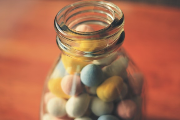 A glass jar containing colorful candy-coated chocolate eggs sits on a warm-toned surface. The jar is partially filled, with the candy displaying pastel shades including yellow, pink, blue, and cream.