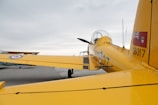 A yellow aircraft is parked on an airfield with visible markings and insignias. The aircraft's tail and wings are prominently featured, displaying detailed emblems and numbers. In the background, another aircraft can be seen, partially obscuring the sky.