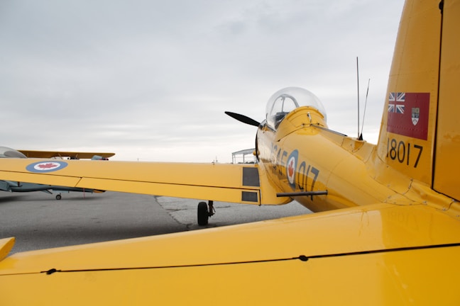 A yellow aircraft is parked on an airfield with visible markings and insignias. The aircraft's tail and wings are prominently featured, displaying detailed emblems and numbers. In the background, another aircraft can be seen, partially obscuring the sky.