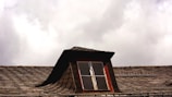 A rooftop with wooden shingles features a small dormer window. The window frame is painted red and is slightly weathered. The sky is overcast with thick clouds, creating a moody atmosphere.
