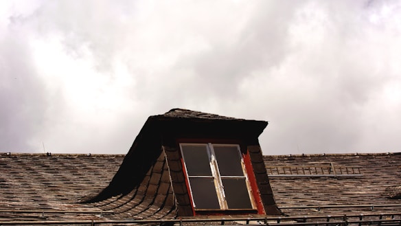 A rooftop with wooden shingles features a small dormer window. The window frame is painted red and is slightly weathered. The sky is overcast with thick clouds, creating a moody atmosphere.