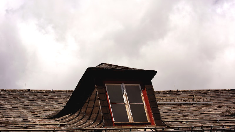 A rooftop with wooden shingles features a small dormer window. The window frame is painted red and is slightly weathered. The sky is overcast with thick clouds, creating a moody atmosphere.