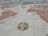 A detailed world map is depicted on a stone or tiled surface. The map includes illustrations of old sailing ships positioned on various parts of the ocean, with a prominent compass rose placed in the lower section of the image. The continents are shaded in a reddish hue, and the map is laid out seemingly on a horizontal surface, like a plaza or square.