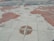 A detailed world map is depicted on a stone or tiled surface. The map includes illustrations of old sailing ships positioned on various parts of the ocean, with a prominent compass rose placed in the lower section of the image. The continents are shaded in a reddish hue, and the map is laid out seemingly on a horizontal surface, like a plaza or square.