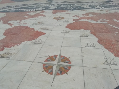 A detailed world map is depicted on a stone or tiled surface. The map includes illustrations of old sailing ships positioned on various parts of the ocean, with a prominent compass rose placed in the lower section of the image. The continents are shaded in a reddish hue, and the map is laid out seemingly on a horizontal surface, like a plaza or square.