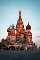 The image features a vibrant and intricately designed building with colorful onion-shaped domes set against a clear sky. People are gathered around, some walking and others taking photos, adding a lively atmosphere to the scene. The architecture is detailed with patterns and a mix of contrasting colors.