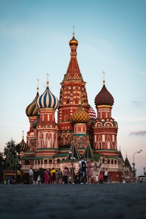 The image features a vibrant and intricately designed building with colorful onion-shaped domes set against a clear sky. People are gathered around, some walking and others taking photos, adding a lively atmosphere to the scene. The architecture is detailed with patterns and a mix of contrasting colors.