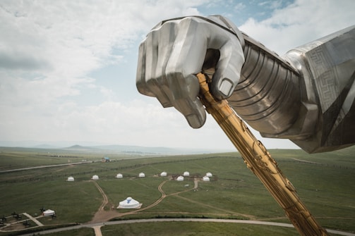 A large metallic structure depicting a hand holding a golden staff dominates the foreground, set against a vast green landscape. In the background, traditional yurt tents are scattered across the open field, creating a stark contrast between modern industrial art and traditional culture. The sky is partly cloudy, adding a sense of openness to the scene.