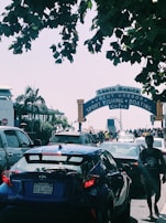 A busy scene at the entrance of the Santa Monica Pier with multiple cars in traffic and several pedestrians walking. Green trees partially overshadow the area. The iconic sign reads 'Santa Monica Yacht Harbor Sport Fishing Boating Cafes.' The Ferris wheel from the amusement park is visible in the background.