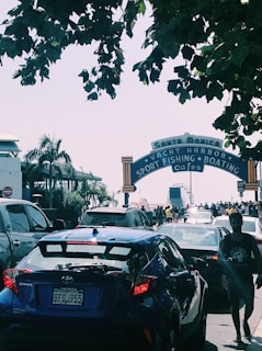 A busy scene at the entrance of the Santa Monica Pier with multiple cars in traffic and several pedestrians walking. Green trees partially overshadow the area. The iconic sign reads 'Santa Monica Yacht Harbor Sport Fishing Boating Cafes.' The Ferris wheel from the amusement park is visible in the background.
