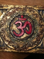 An ornate decorative piece featuring the Om symbol in red at the center, surrounded by intricate patterns in gold, green, and brown. The design is set against a textured golden background with a circular frame that adds an antiquated look.