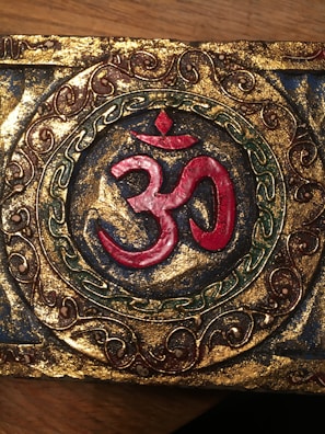 An ornate decorative piece featuring the Om symbol in red at the center, surrounded by intricate patterns in gold, green, and brown. The design is set against a textured golden background with a circular frame that adds an antiquated look.