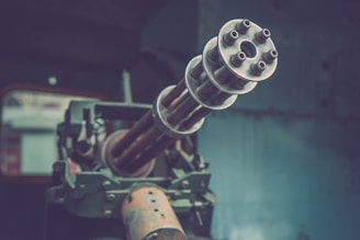 Close-up view of a multi-barreled rotary gun pointing outward. The gun appears to be mounted on a vehicle or a fixed structure. The background is out of focus, indicating an industrial or military setting.