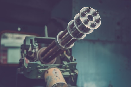 Close-up view of a multi-barreled rotary gun pointing outward. The gun appears to be mounted on a vehicle or a fixed structure. The background is out of focus, indicating an industrial or military setting.