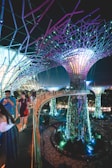 A futuristic scene with large, illuminated tree-like structures surrounded by vibrant vegetation. People walk along an elevated pathway that curves through the scene, with lights casting colorful glows throughout the area. The overall environment is a blend of nature and technology, with intricate designs and immersive lighting.