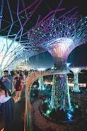 A futuristic scene with large, illuminated tree-like structures surrounded by vibrant vegetation. People walk along an elevated pathway that curves through the scene, with lights casting colorful glows throughout the area. The overall environment is a blend of nature and technology, with intricate designs and immersive lighting.
