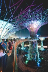 A futuristic scene with large, illuminated tree-like structures surrounded by vibrant vegetation. People walk along an elevated pathway that curves through the scene, with lights casting colorful glows throughout the area. The overall environment is a blend of nature and technology, with intricate designs and immersive lighting.