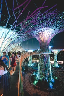 A futuristic scene with large, illuminated tree-like structures surrounded by vibrant vegetation. People walk along an elevated pathway that curves through the scene, with lights casting colorful glows throughout the area. The overall environment is a blend of nature and technology, with intricate designs and immersive lighting.