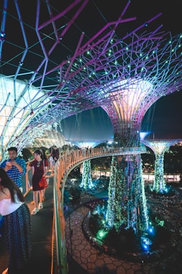 A futuristic scene with large, illuminated tree-like structures surrounded by vibrant vegetation. People walk along an elevated pathway that curves through the scene, with lights casting colorful glows throughout the area. The overall environment is a blend of nature and technology, with intricate designs and immersive lighting.
