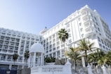 A grand, multi-story white hotel with numerous balconies and architectural details is surrounded by palm trees. An ornate white gazebo stands in the foreground, and the clear blue sky enhances the serene ambiance.