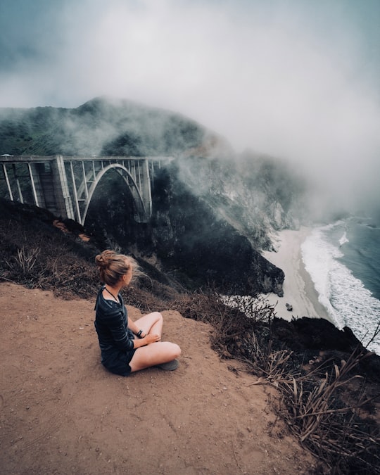 woman in black top sitting beside of mountain cliff during cloudy sky in Bixby Creek Bridge United States