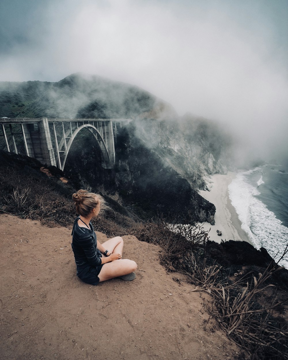 woman in black top sitting beside of mountain cliff during cloudy sky