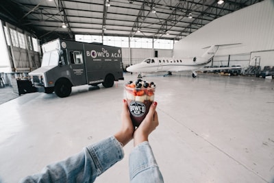 Inside a spacious hangar, a food truck labeled 'BOWL'D ACAI' is parked on the left, while a small private jet is positioned towards the back. In the foreground, two hands hold a colorful acai bowl topped with fruits. The hangar is well-lit with natural and artificial lighting, and the floor is clean and spacious.