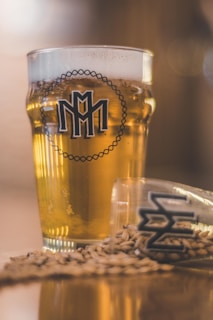 A full pint glass containing a golden-hued beverage, possibly beer, prominently displaying a logo on its front. Beside it, a smaller glass lies on its side with similar branding, and grain or barley is scattered around on the surface, contributing to a rustic and artisanal impression.