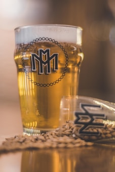 A full pint glass containing a golden-hued beverage, possibly beer, prominently displaying a logo on its front. Beside it, a smaller glass lies on its side with similar branding, and grain or barley is scattered around on the surface, contributing to a rustic and artisanal impression.