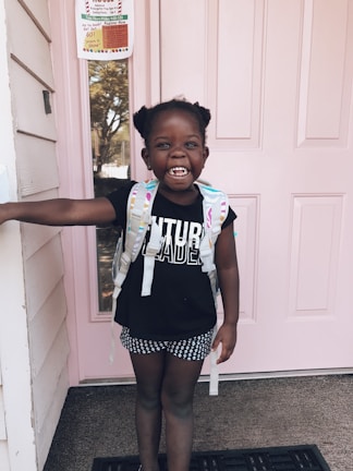 A young child stands in front of a pink door wearing a black shirt with 'Future Leader' written on it, polka-dot shorts, and a backpack. The child has a big smile and has one arm extended to touch the doorframe. There is a notice or poster visible on the door behind the child.