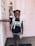 A young child stands in front of a pink door wearing a black shirt with 'Future Leader' written on it, polka-dot shorts, and a backpack. The child has a big smile and has one arm extended to touch the doorframe. There is a notice or poster visible on the door behind the child.