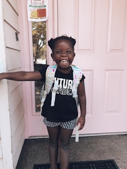A young child stands in front of a pink door wearing a black shirt with 'Future Leader' written on it, polka-dot shorts, and a backpack. The child has a big smile and has one arm extended to touch the doorframe. There is a notice or poster visible on the door behind the child.