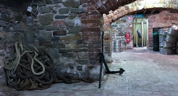 A rustic basement with stone and brick walls, featuring a pile of coiled ropes on the floor. There is an anchor leaning against the wall, and several metal barrels are stacked in the background. The area is dimly lit, highlighting an arched brick ceiling and a narrow corridor leading to an exit.