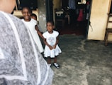 Two young children, dressed in white, stand in a simple, rustic setting with concrete floors and unpainted walls. The children are looking towards the camera, with one smiling and holding hands with the other. The space appears to be a modest indoor environment with a wooden chair and a few items visible in the background.
