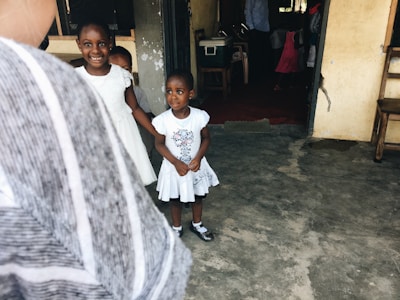 Two young children, dressed in white, stand in a simple, rustic setting with concrete floors and unpainted walls. The children are looking towards the camera, with one smiling and holding hands with the other. The space appears to be a modest indoor environment with a wooden chair and a few items visible in the background.
