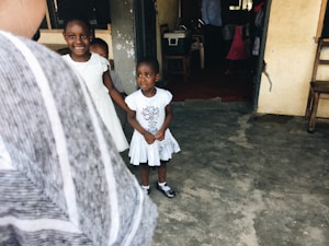 Two young children, dressed in white, stand in a simple, rustic setting with concrete floors and unpainted walls. The children are looking towards the camera, with one smiling and holding hands with the other. The space appears to be a modest indoor environment with a wooden chair and a few items visible in the background.