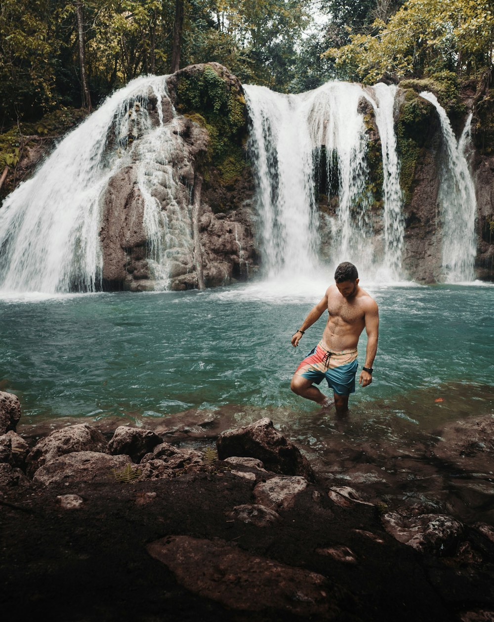 man in blue board shorts standing on rocks on wateralls side