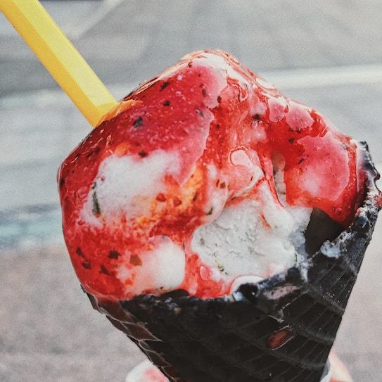 A close-up of a melting ice cream with vibrant red syrup drizzled on top, held in a black waffle cone with a yellow spoon sticking out.