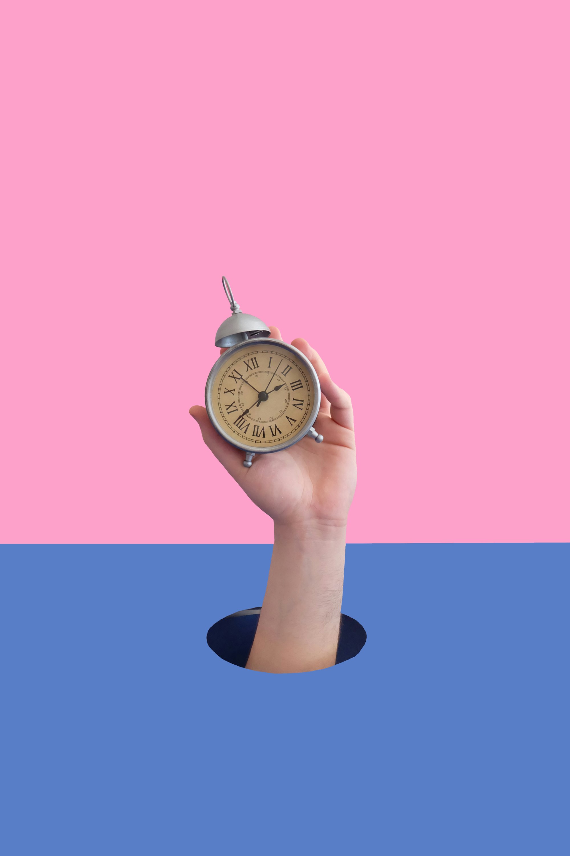 Hand holding an alarm clock, reaching through a whole with pink and lilac background.