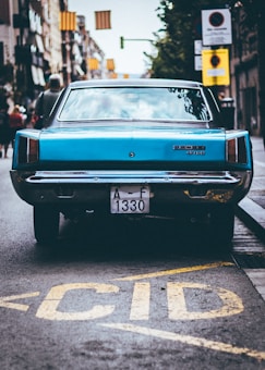 A classic blue car with a 'Dodge 3700' badge is parked on a city street. The vehicle's rear view is prominent, and its license plate reads 'A-F 1330'. The car is positioned near the curb with colorful banners visible above, and blurred pedestrians are seen walking in the background.