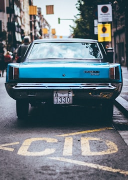 A classic blue car with a 'Dodge 3700' badge is parked on a city street. The vehicle's rear view is prominent, and its license plate reads 'A-F 1330'. The car is positioned near the curb with colorful banners visible above, and blurred pedestrians are seen walking in the background.
