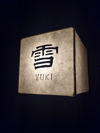 A cube-shaped lamp is softly illuminated with a textured surface. It features a large Kanji character above the word 'YUKI' on a glowing backdrop, set against a dark background.