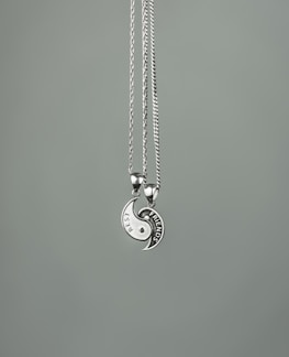 Two necklaces with circular pendants form a yin-yang shape. Each half has an engraved design, one labeled 'BEST' and the other 'FRIENDS'. The pendants are metallic with a shiny finish, suspended on thin chains against a plain gray background.