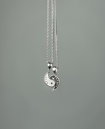 Two necklaces with circular pendants form a yin-yang shape. Each half has an engraved design, one labeled 'BEST' and the other 'FRIENDS'. The pendants are metallic with a shiny finish, suspended on thin chains against a plain gray background.
