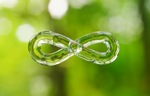 A clear, glass-like infinity symbol is suspended in the foreground against a blurred, vibrant green background, suggesting nature or foliage.
