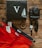 A collection of items laid out on a wooden surface. The items include three pairs of pants in red, navy, and black from the brand Cove Five, a black box with a large white letter V and a sailboat illustration, a wooden bead bracelet, a black watch with a white face, black sunglasses, and a Sony Alpha camera.
